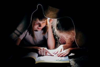 Two young girls read a book via a flashlight while under a blanket