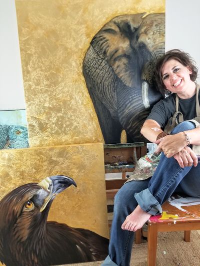 Artists Hilary Clement sits in her studio, barefoot and wearing he painter's apron, in front of original paintings of an elephant and an eagle.
